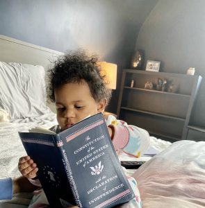 A toddler, Stephanie Branton's granddaughter, sits on a bed holding a copy of the United States Constitution
