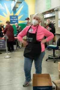A woman in a pink shirt, mask and apron that says "friends of CALS volunteer" stands with her hands on her hips in a sassy pose.