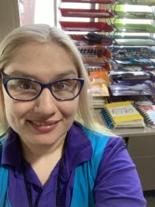 A woman wearing glasses and a blue and purple shirt, in front of a colorful stack of shelves