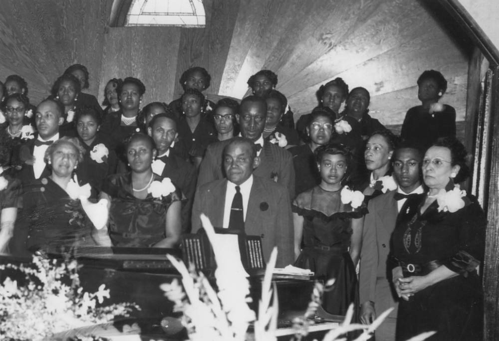 Group of black men and women in dark suits and dresses with women wearing white flowers on shoulder.
